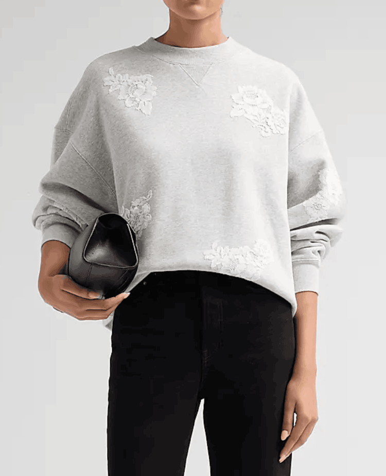 A model wearing a light gray crewneck sweatshirt with white floral embroidery on the chest, paired with black pants and holding a black clutch bag against a light gray background.